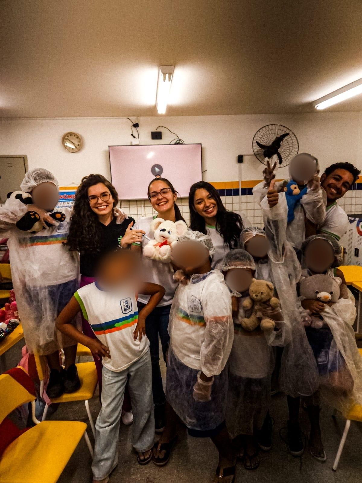 Crianças de touca e jaleco levantando os ursinhos de pelúcia, ao lado das integrantes da equipe, no fim do evento